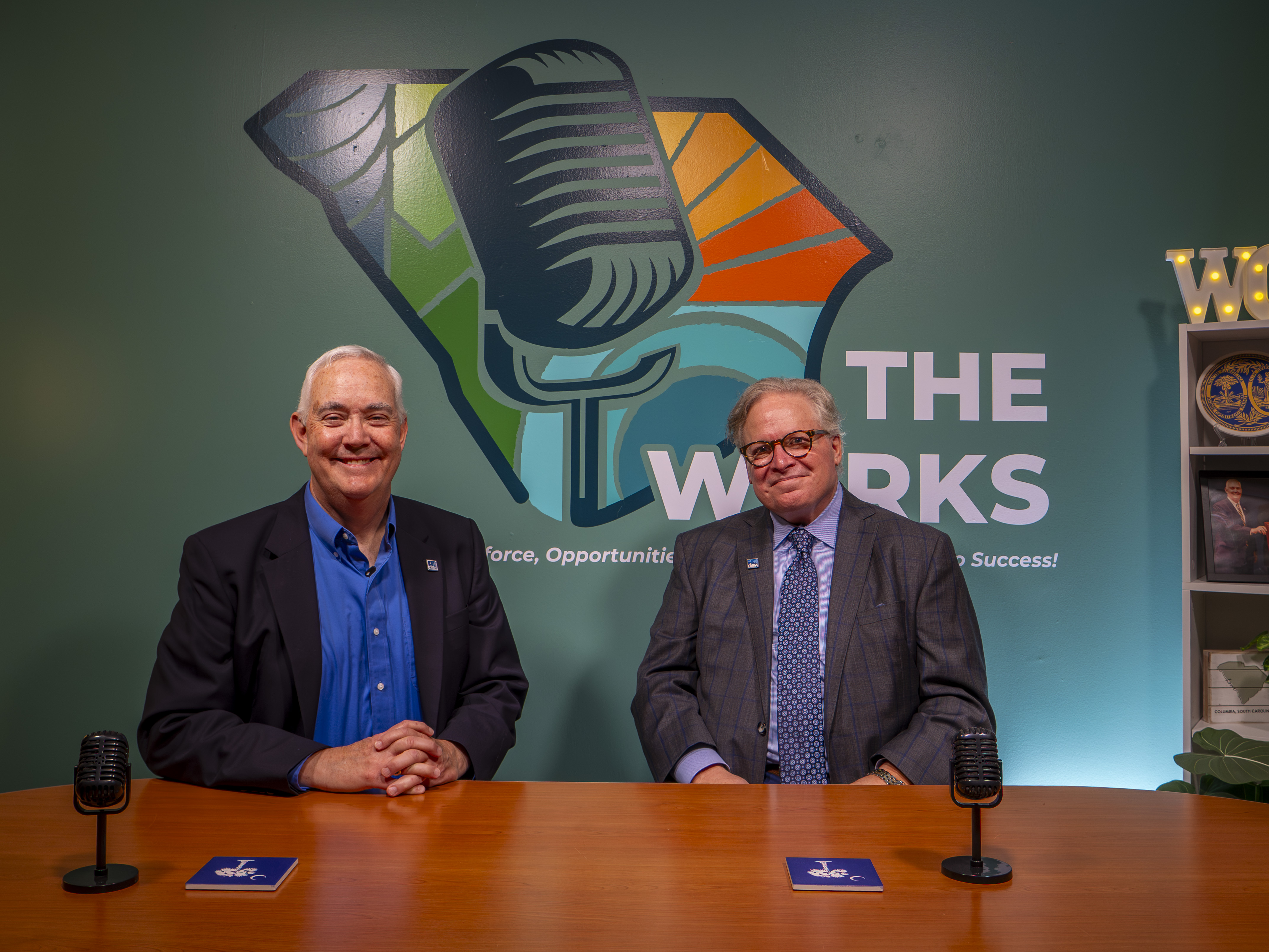 Two men in suits sit at a table in front of microphones.