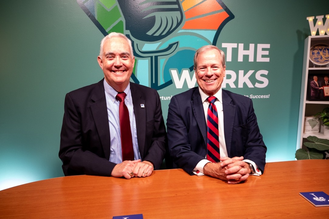 Two men in suits are seated at a table with their hands clasped. Both men are smiling. There is a The Works logo behind their heads on the wall.