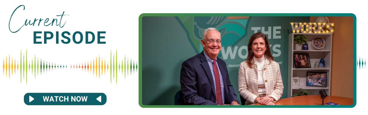 A man and a woman sit next to each other at a desk. Both are smiling. There is a microphone in front of them.
