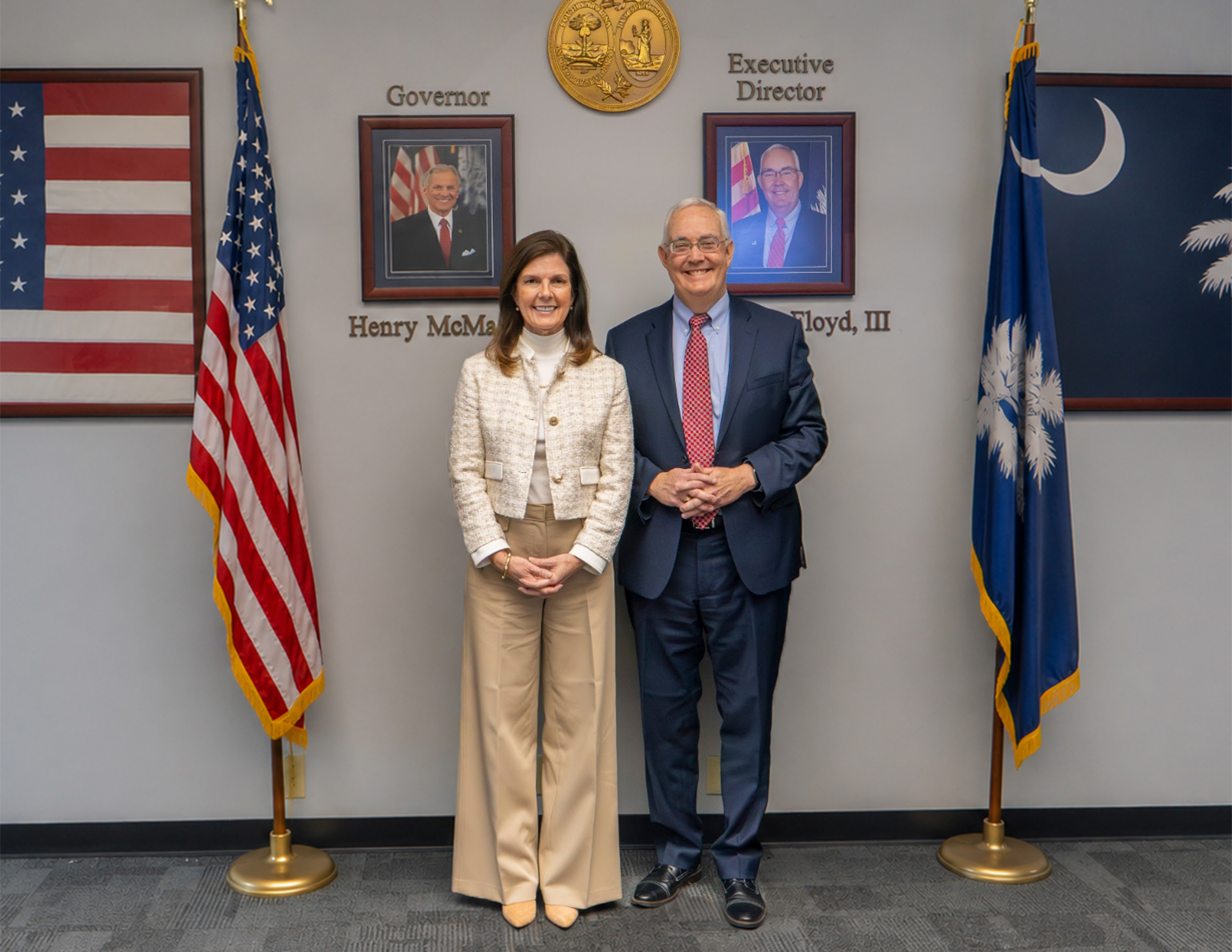 A woman in a tan and white suit is standing next to a man in a navy suit. Both are smiling. The South Carolina State Flag and the USA flag are on either side.