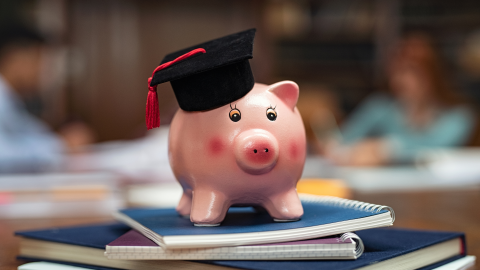 piggy bank wearing grad cap on top of stack of books in library setting with focal blurred background
